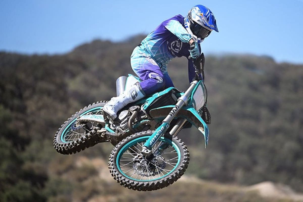 Rider in purple and teal gear whipping a teal Kove MX450 sideways in mid-air above a hillside motocross track