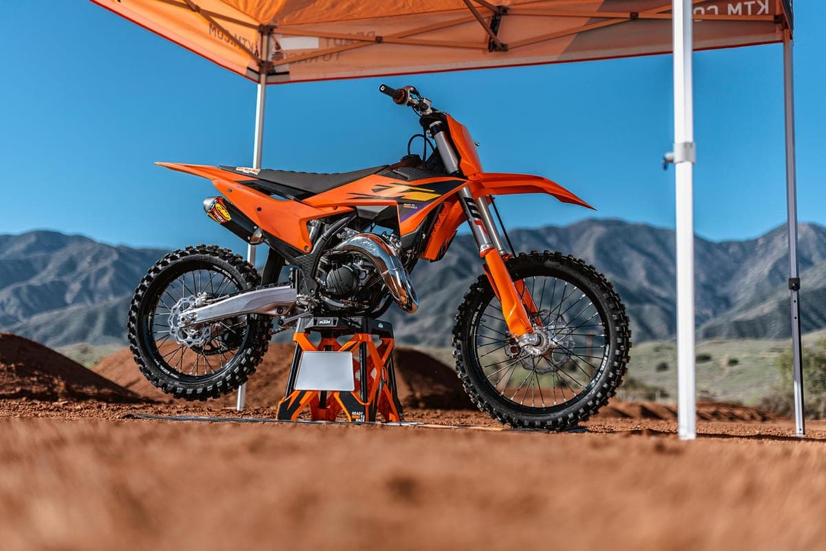 KTM 125 SX parked on an orange KTM stand under a pop-up canopy at a dirt track, mountains and blue sky in the background