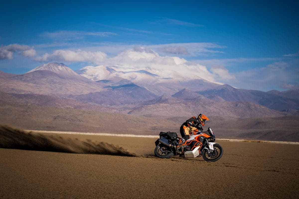 Rider in orange KTM gear carving through a desert dry lakebed on the Super Adventure R, kicking up a long trail of dust with snow-capped mountains behind