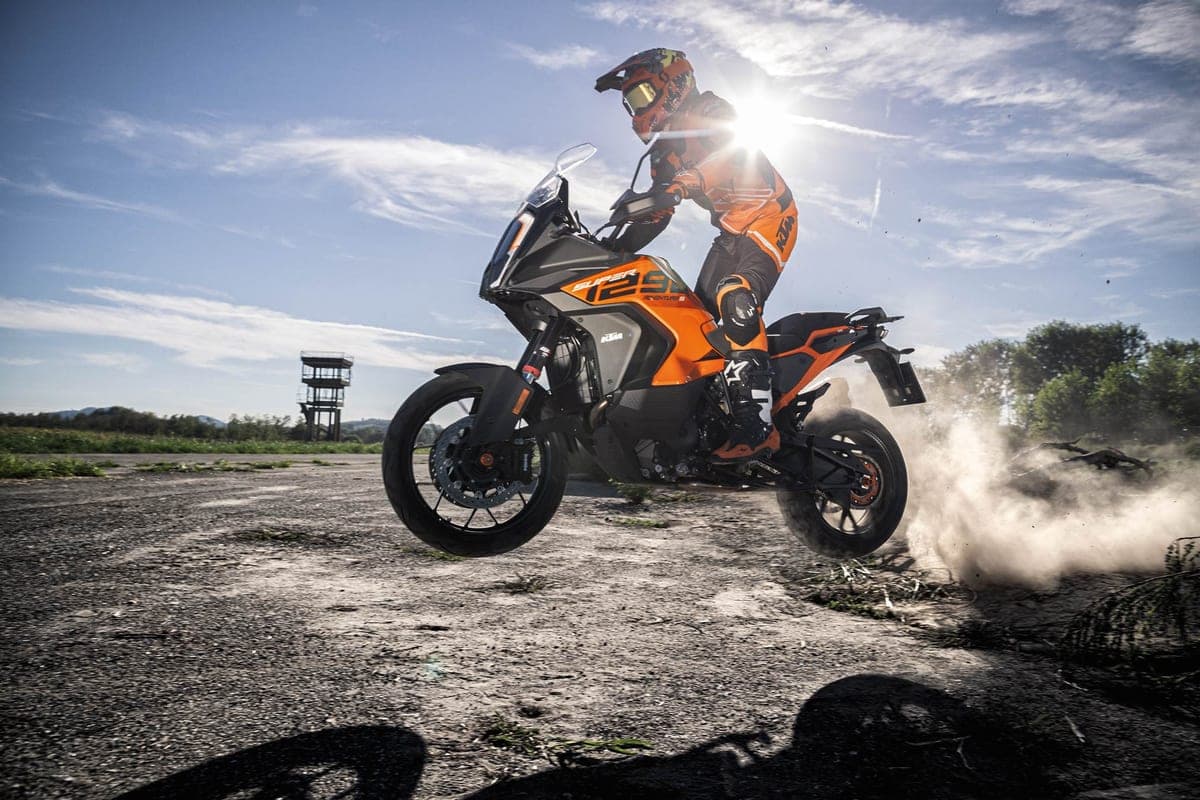 Rider in orange riding gear launching a KTM adventure bike off a jump on tarmac, dust exploding behind the rear wheel under bright sun flare