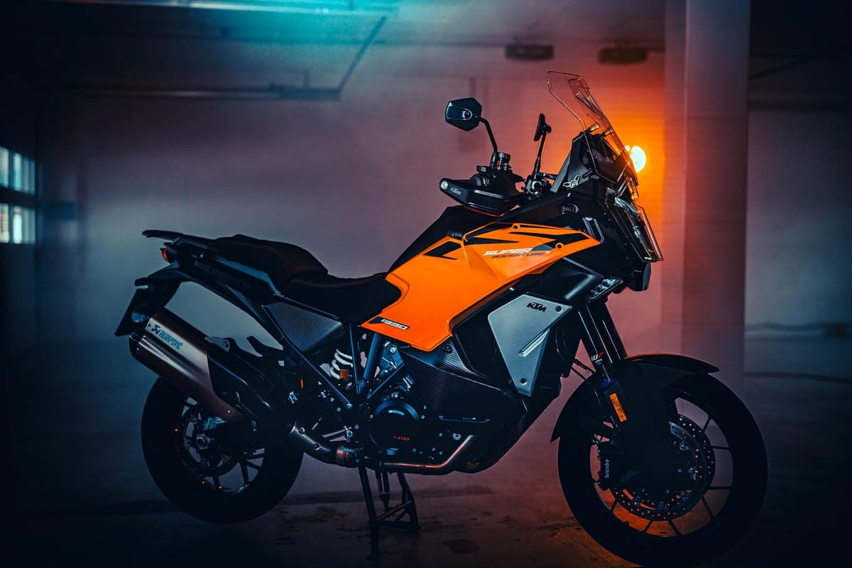 KTM 1390 Super Adventure S EVO left-side studio shot in a dim garage with orange accent lighting, showing the Akrapovic exhaust and beak-style front fender