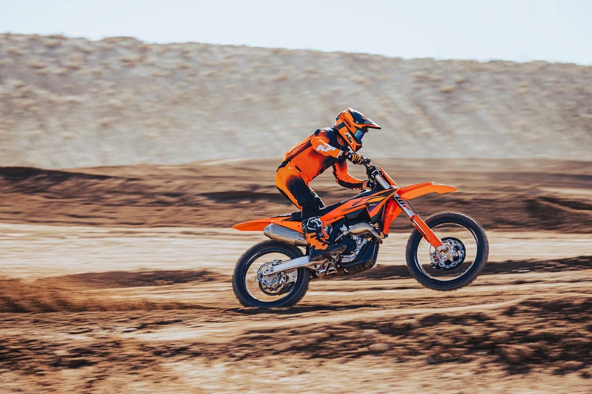 KTM 250 SX-F rider in orange and black gear launching off a jump on a sandy motocross track, front wheel lofted skyward