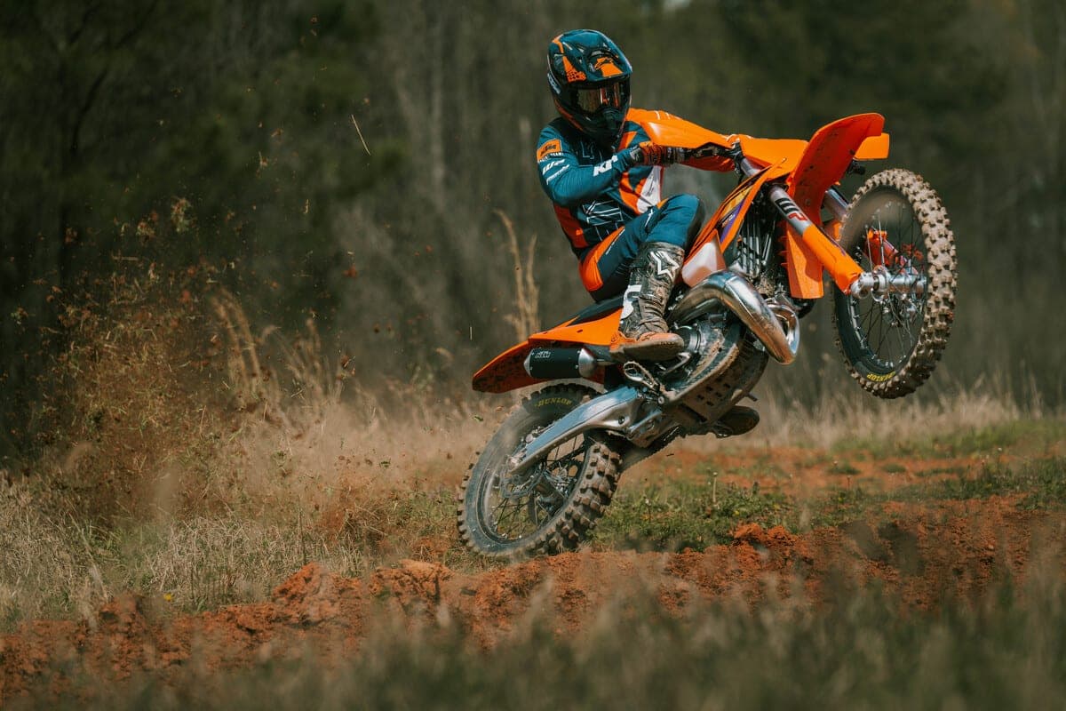 Rider in orange and teal KTM gear popping a wheelie on a KTM 250 XC through a red-dirt trail, kicking up dust and roost from the rear tire