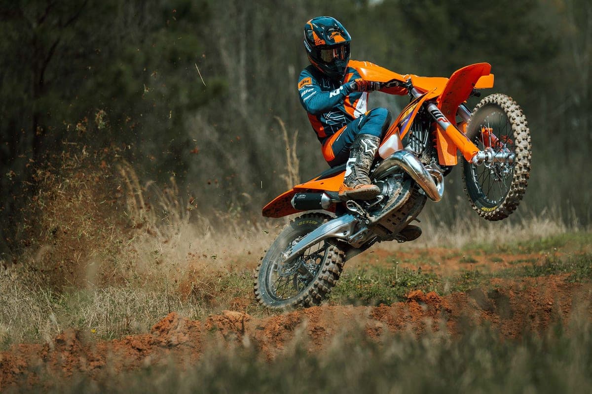 Rider in orange and blue KTM gear whipping the 250 XC-F front wheel up over a red-clay trail berm, kicking up a spray of dirt