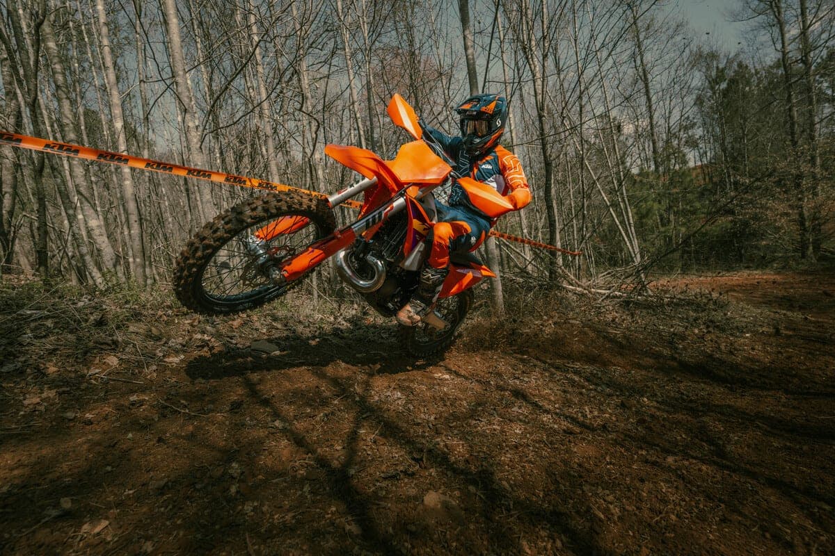 Rider in orange and blue gear popping a wheelie on a 2027 KTM 250 XC-W through a wooded dirt trail lined with tape