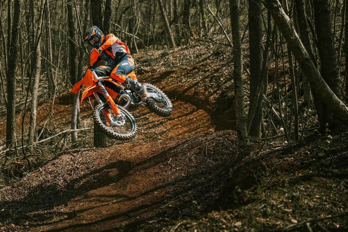 Rider in orange and grey KTM gear launching a 300 XC off a dirt jump on a shaded forest enduro trail