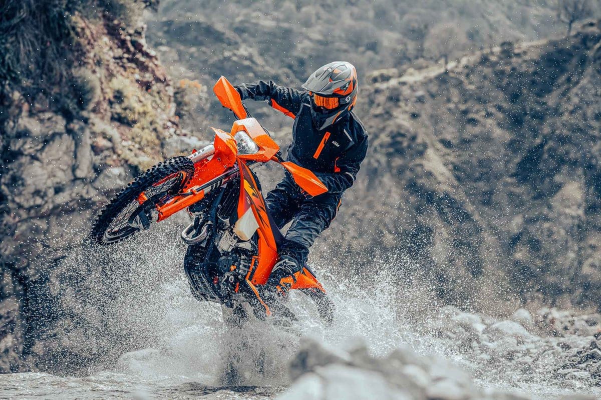 Rider in black and orange gear splashing a KTM 350 EXC-F through a rocky stream crossing, front wheel lifted over the water