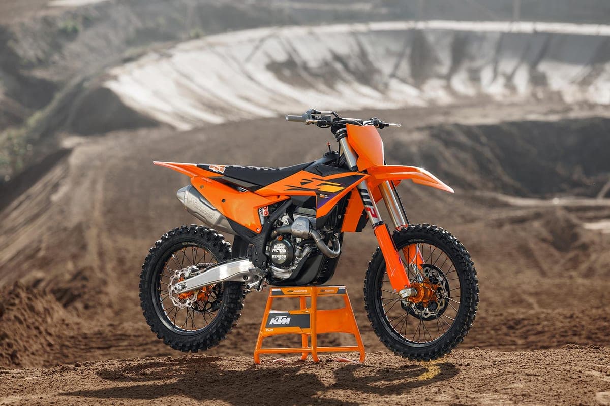 KTM 350 SX-F parked on an orange KTM stand at a dirt motocross track, side profile against sculpted sand berms and hillside terrain