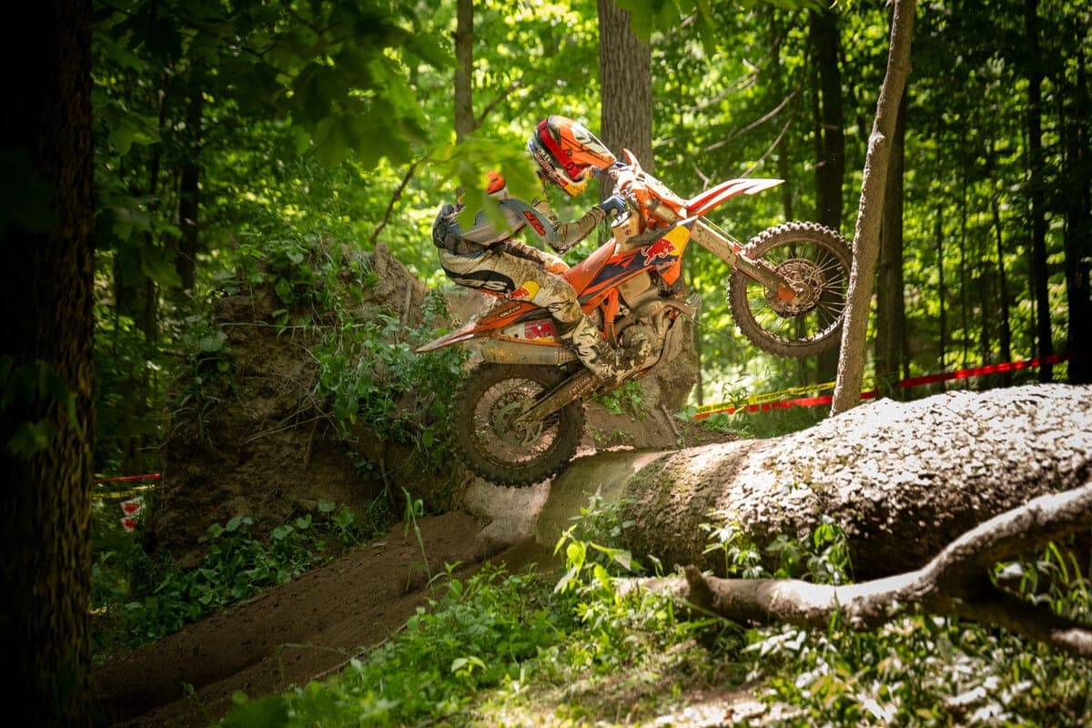 Rider in muddy gear atop a KTM 350 XC-F Factory Edition, powering over a log obstacle on a shaded forest enduro trail