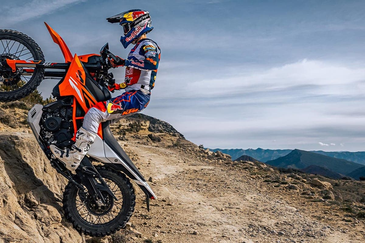 Rider in Red Bull KTM gear hoisting the KTM 390 Enduro R up a rocky hillside, front wheel lifted off a steep dirt ledge