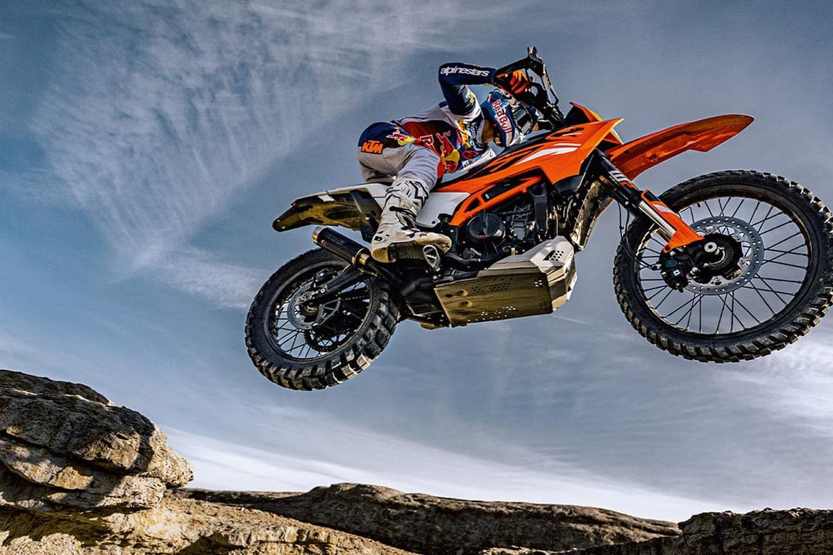 KTM 390 Enduro R airborne over rocks with a rider in white and orange gear tucked low, bash plate and single-sided exhaust visible mid-jump