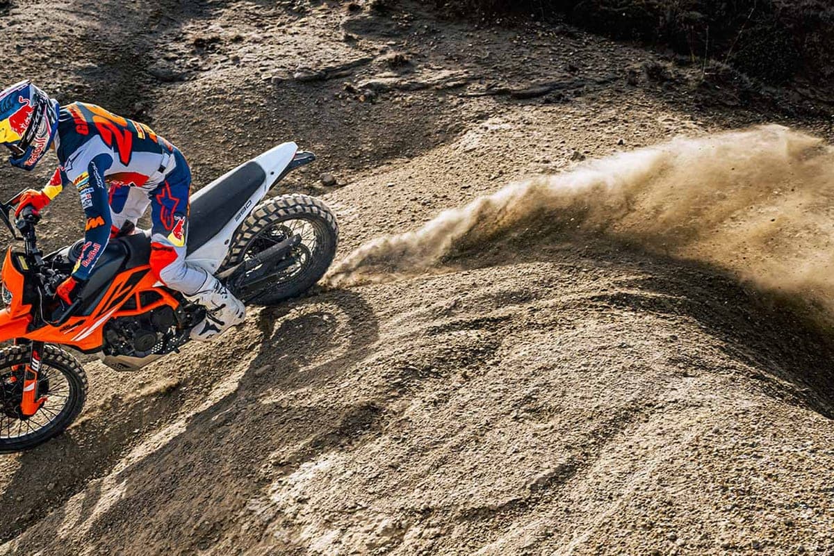 Aerial view of a rider carving the orange and white 390 Enduro R through a dirt berm, rear tire throwing up a wide roost of dust