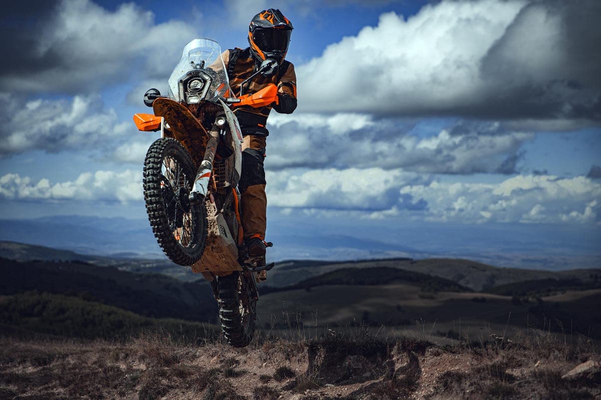 Rider in orange and black gear popping a wheelie on a muddy KTM 390 Adventure R against a cloudy mountain skyline