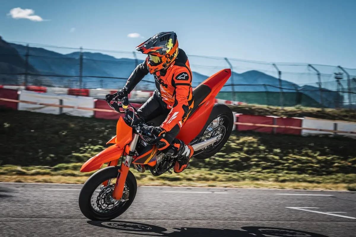 Rider in orange and black Alpinestars gear popping a wheelie on a KTM 450 SMR down a paved race track with mountains behind