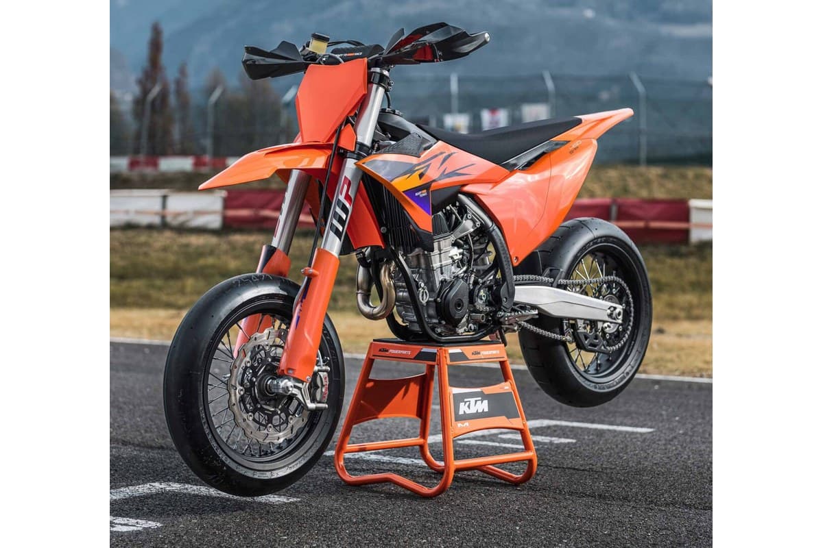 KTM 450 SMR rear three-quarter view on a paddock stand at a track, showing the exposed engine, chain, WP shock and orange bodywork graphics