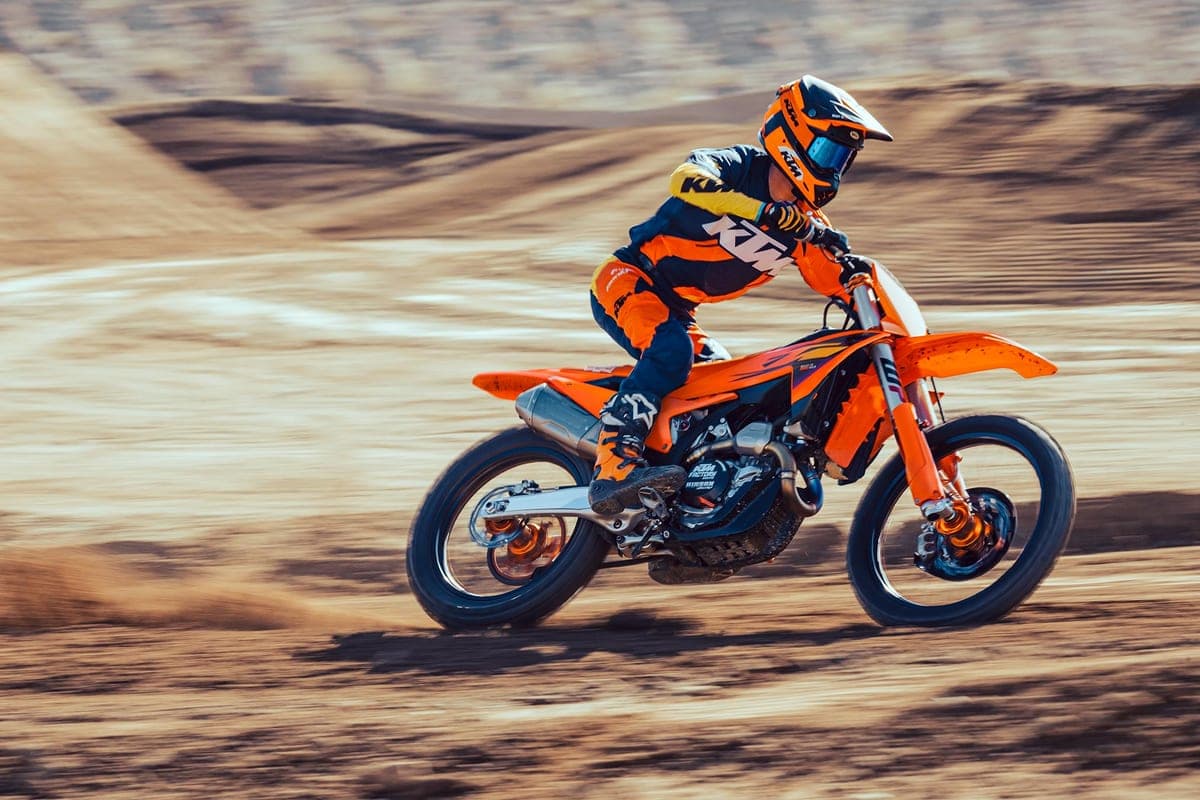 Rider in orange and black KTM gear jumping a bermed motocross section on the KTM 450 SX-F, orange fork legs and rear shock visible