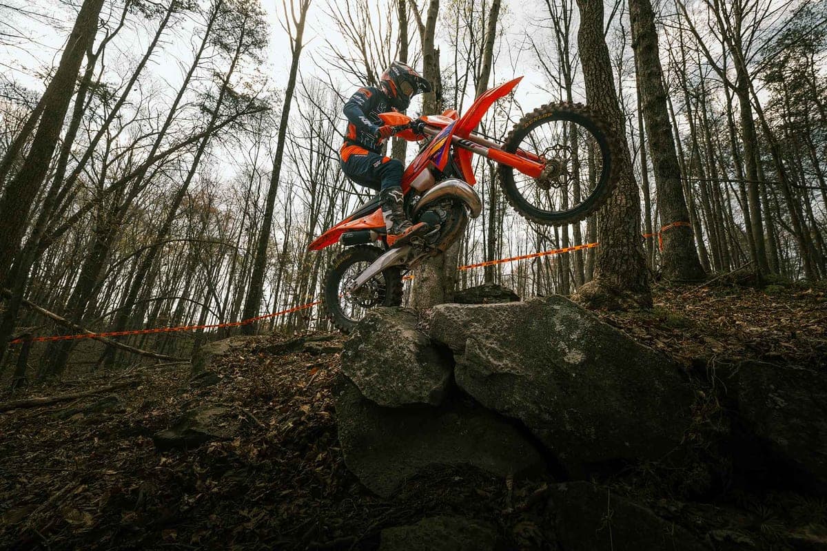Rider in orange KTM gear lofting a 450 XC-F front wheel over a rocky ledge on a wooded enduro trail