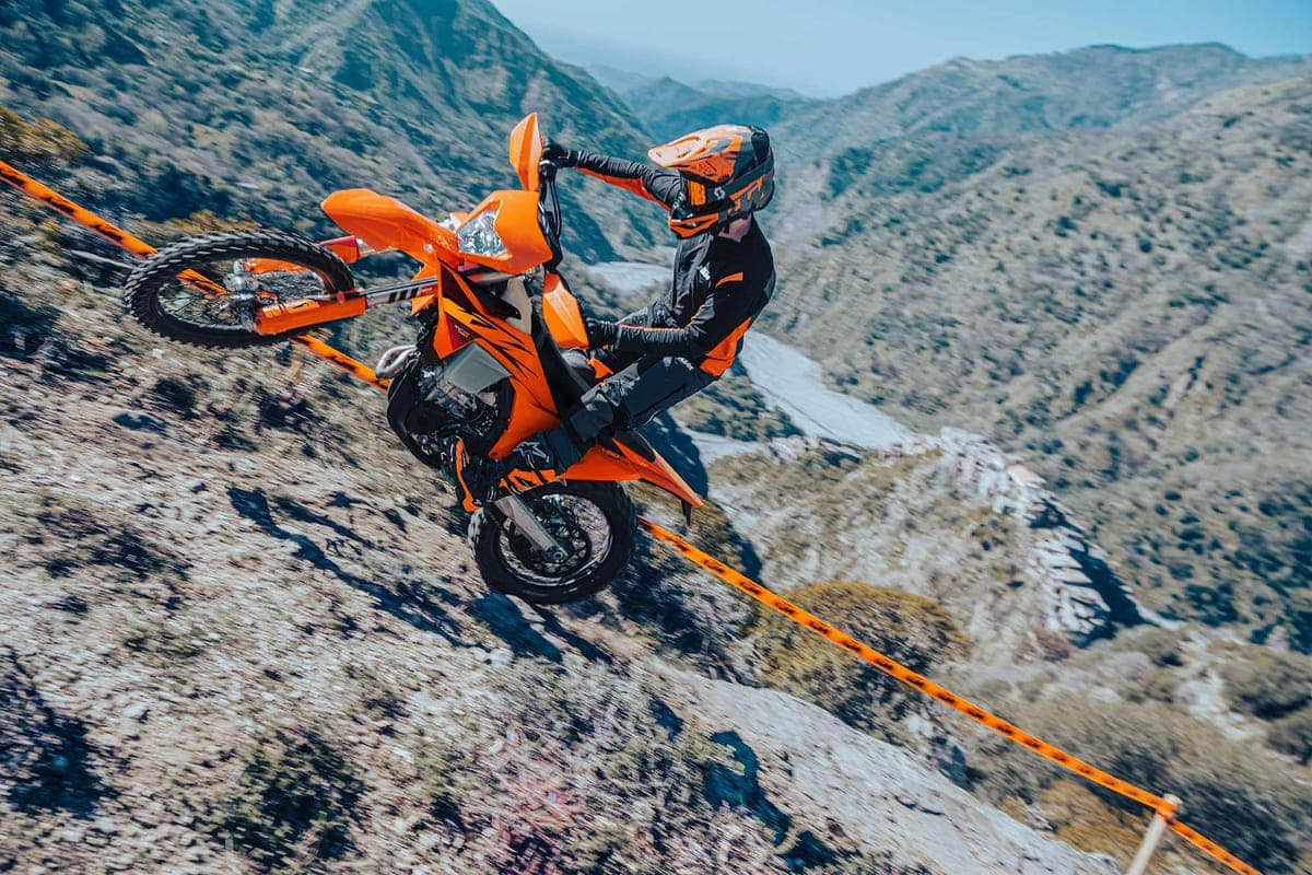 Rider in black and orange gear popping a wheelie on a KTM 450 XCF-W along a steep, rocky mountainside trail marked by orange tape