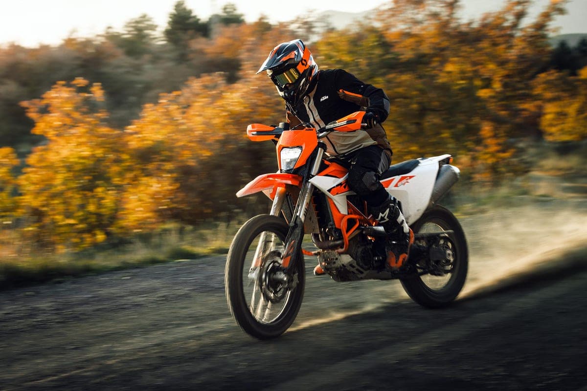 KTM 690 Enduro R in orange and white, front three-quarter action shot with a rider in black and tan gear catching air on a gravel road amid autumn trees