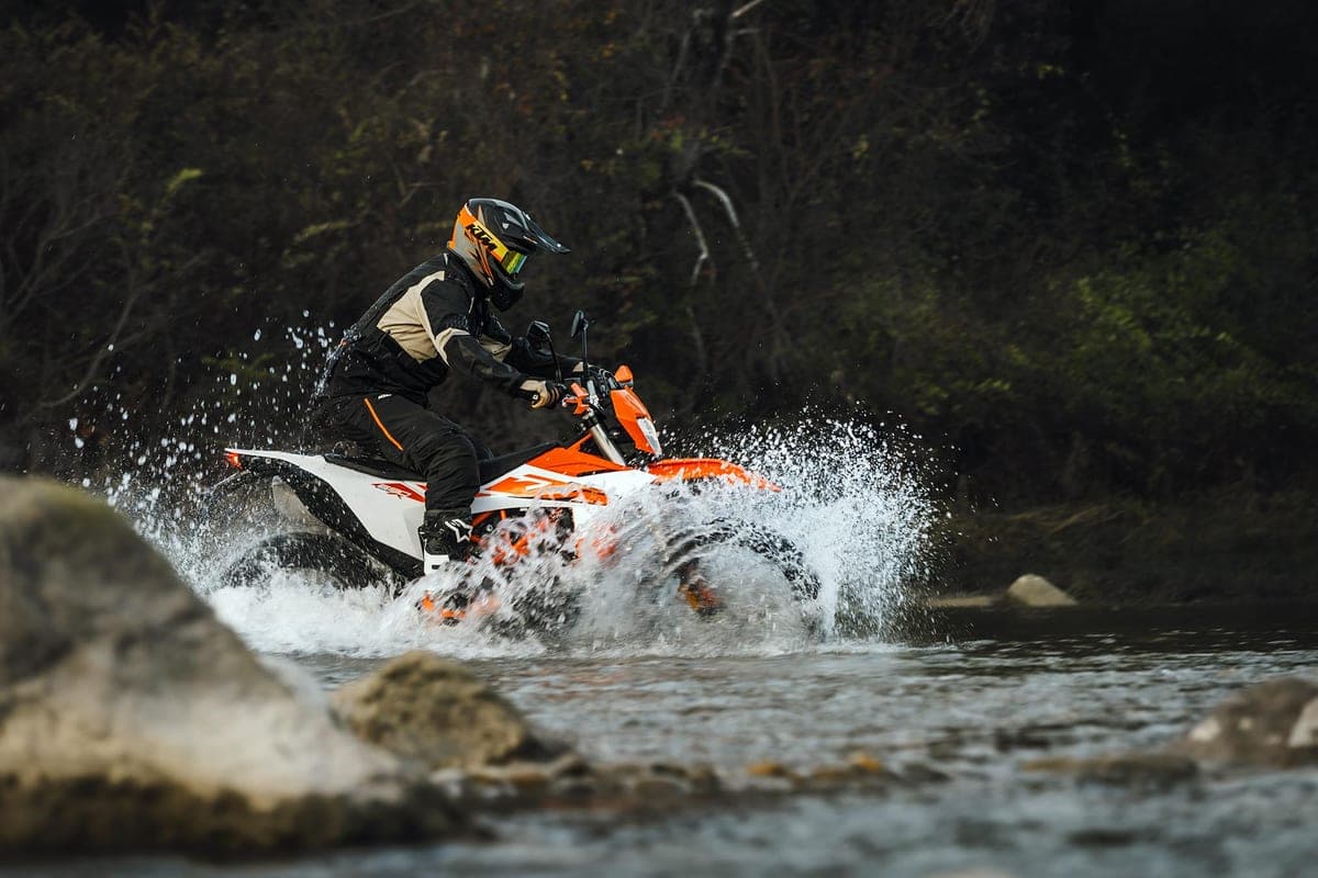 KTM 690 Enduro R in white bodywork, side-on action shot fording a shallow river with a rider in dark gear as spray kicks up around the front wheel