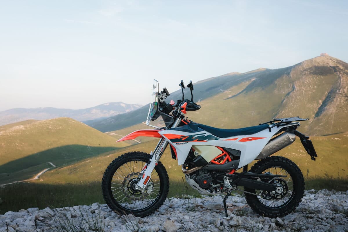KTM 690 Rally parked on a rocky mountain ridge at dusk, right-side profile with green hills and a peak behind it