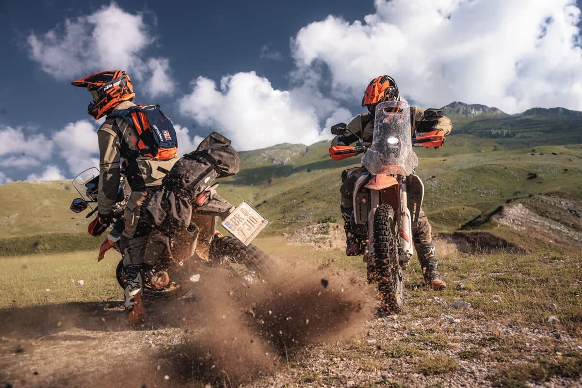 Two riders on KTM 690 Rallys accelerating away up a grassy hillside, throwing loose dirt behind the rear wheels
