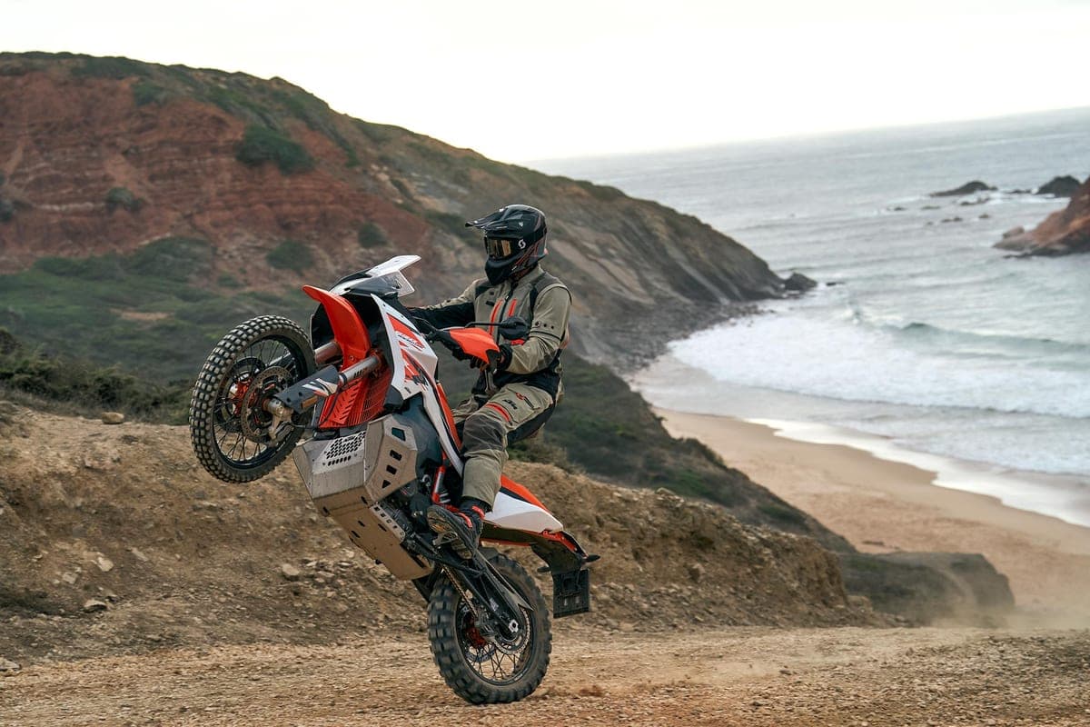 Rider in olive adventure gear popping a wheelie on a KTM 890 Adventure R along a coastal dirt trail above the ocean