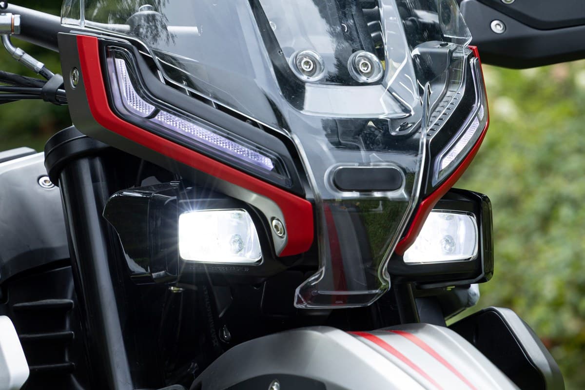 Close-up of the Vettore 450's LED headlight cluster with red trim surrounds and windscreen, set against a blurred green outdoor background