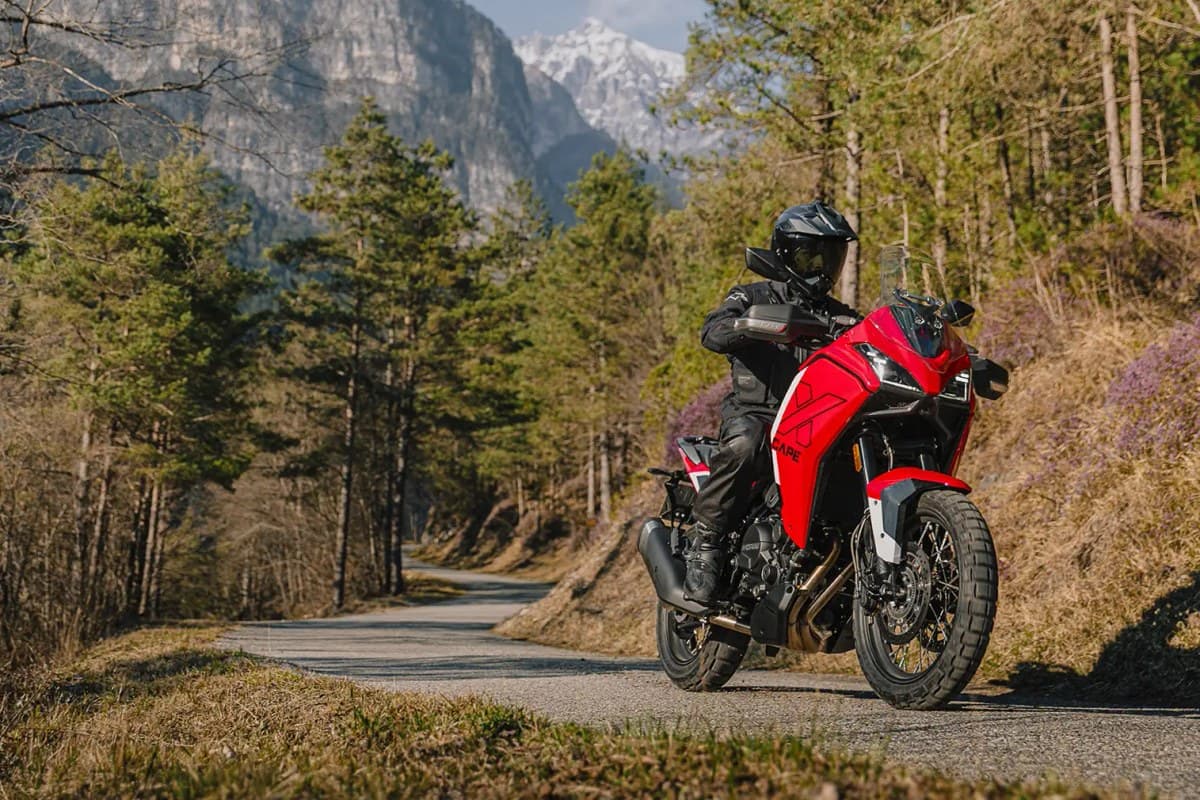 Rider in black gear on a red Moto Morini X-Cape 700, cresting a mountain road lined with pines below snow-capped peaks