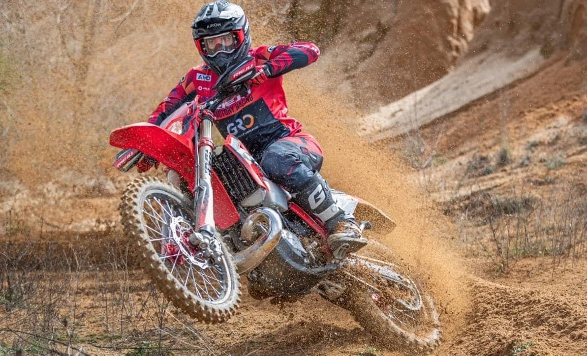 Rider in red Rieju factory racing gear leaning the MR Pro 300i through a sandy turn, roosting a spray of dirt behind