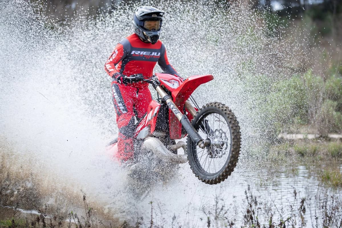 Rider in red and grey Rieju gear blasting the MR Racing 300i through a water crossing, front wheel lofted and spraying a wide arc of spray