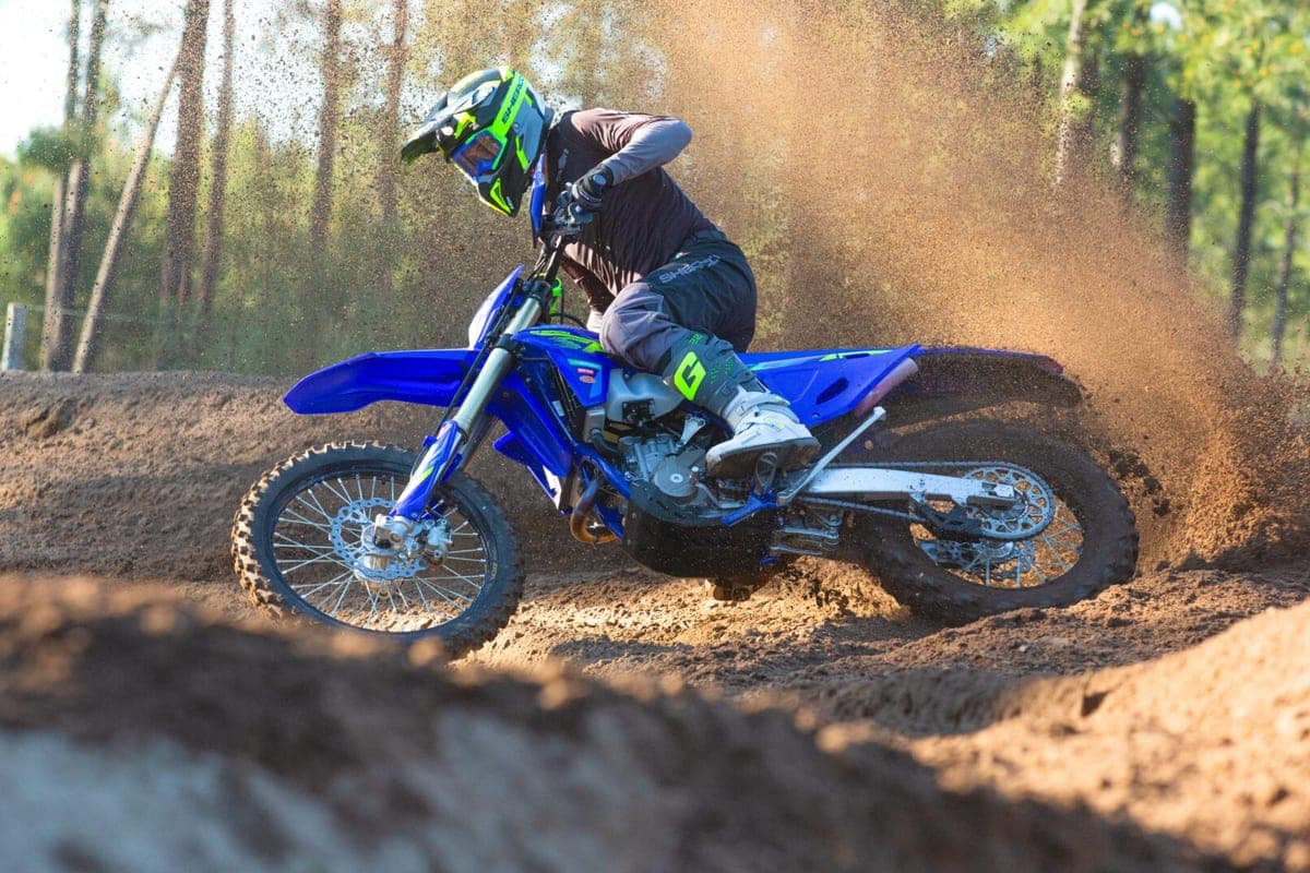 Rider in grey and black gear powering a blue Sherco 450 SEF Factory over a motocross berm, throwing a wall of dirt behind the rear wheel