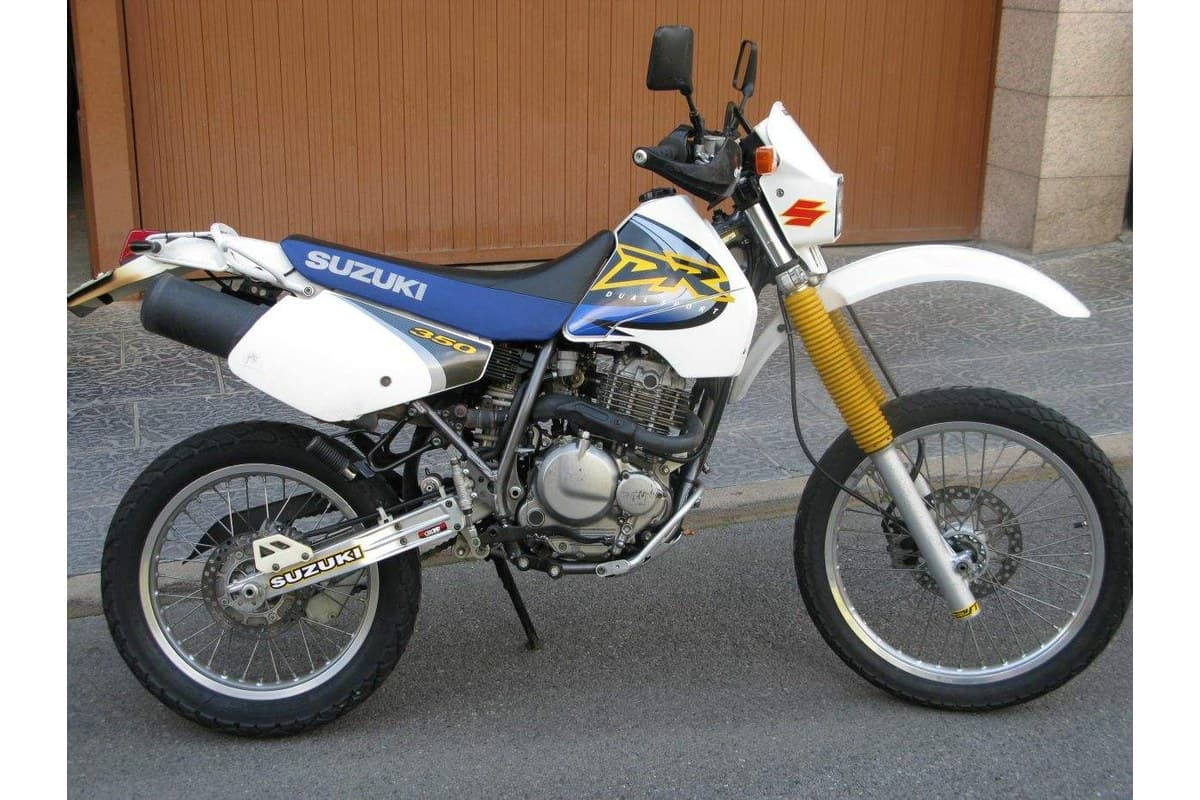Suzuki DR350SE in white and blue livery, left-side view parked outdoors on a driveway with a wood-paneled garage door behind it, gold forks and beak-style front fender