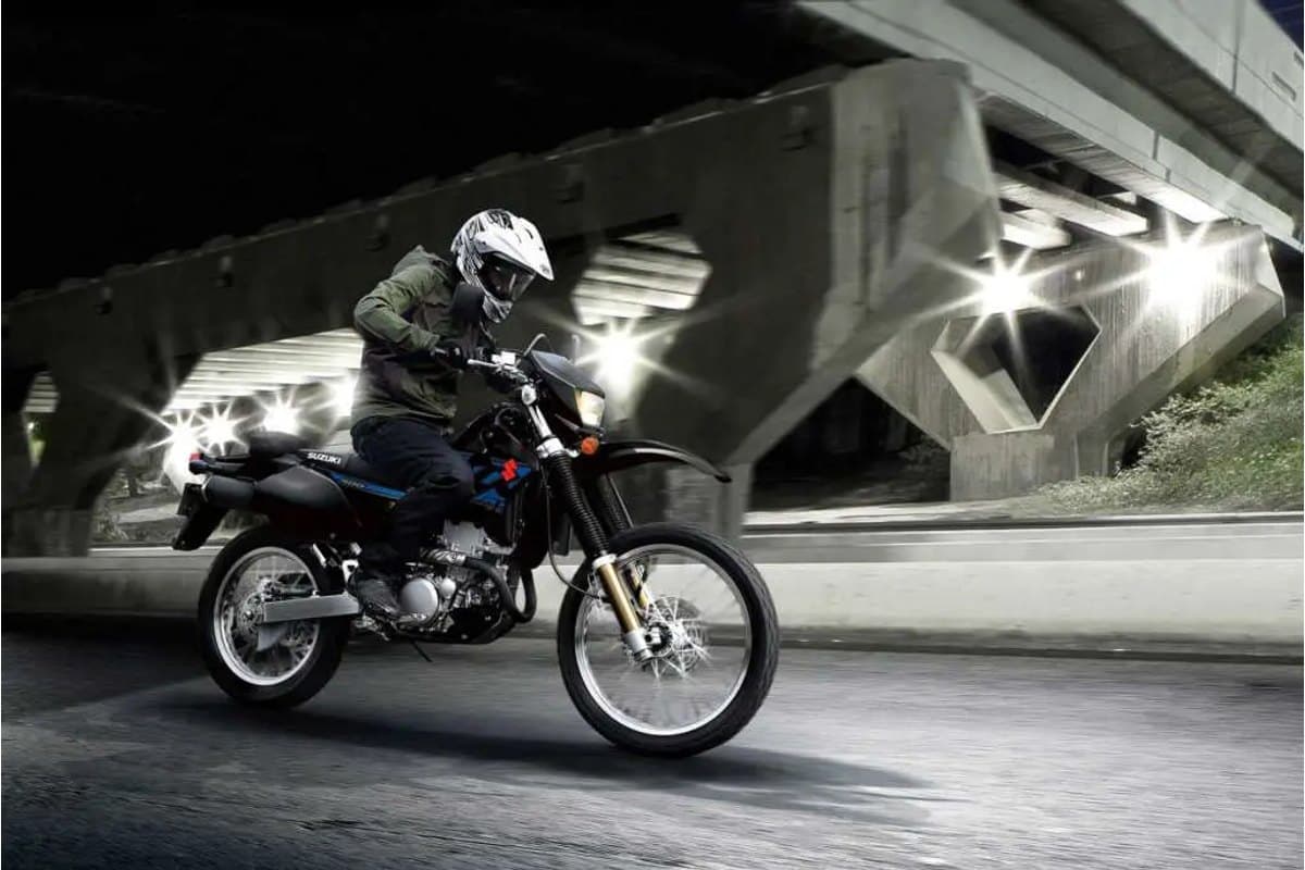 Rider in green jacket and white helmet riding a black DR-Z400S at night under an overpass, headlight beams cutting through the dark roadway