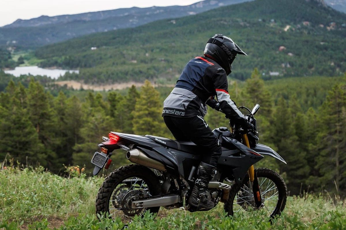 Rider in Alpinestars gear pausing on a Suzuki DR-Z4S atop a grassy overlook, forested mountains and a distant lake behind