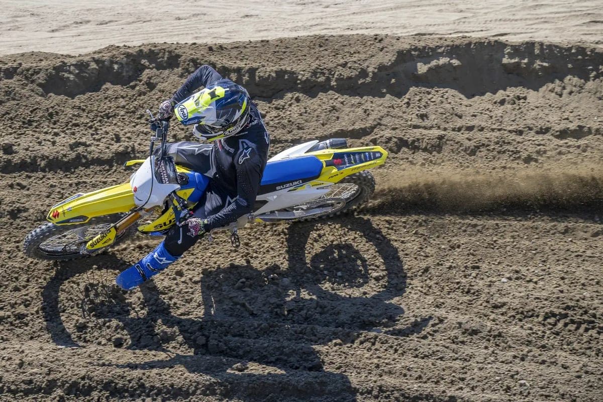 Rider in blue gear cornering a yellow Suzuki RM-Z250 through a sandy motocross berm, spraying a roost of dirt