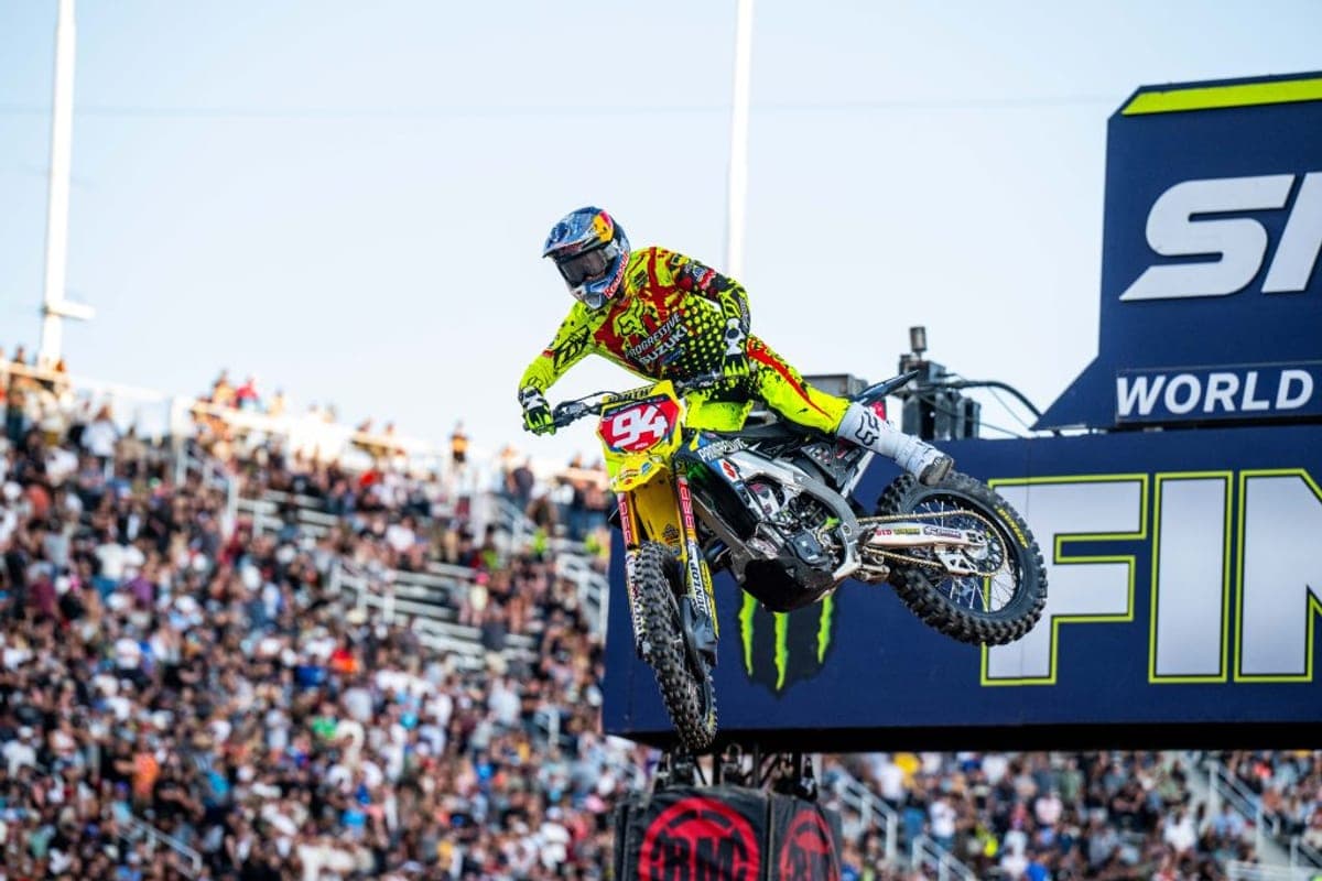 Rider in neon yellow Suzuki race gear whipping the RM-Z450 off a supercross finish-line jump in front of a packed stadium crowd