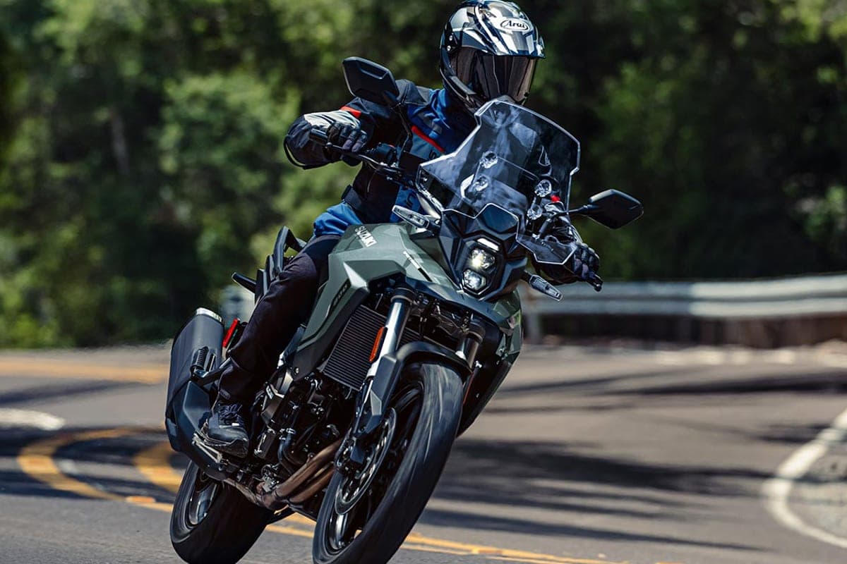 Rider in blue jacket and Arai helmet leaning a matte olive-green Suzuki V-Strom 800 through a wooded mountain curve with the front wheel lifting