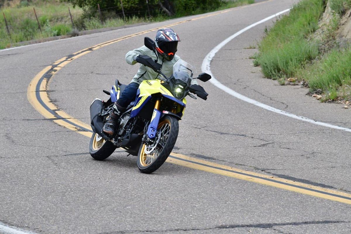 Rider in gear leaning a yellow and blue Suzuki V-Strom 800DE through a winding mountain road, front three-quarter action shot