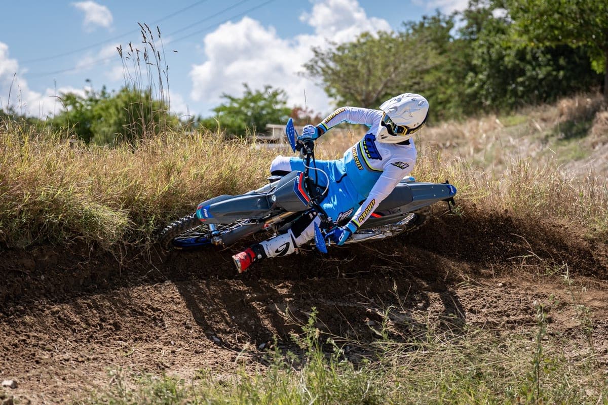 Rider in blue and white gear leaning a grey TM MX 125 2T through a dirt berm, kicking up a spray of loose soil on a motocross track
