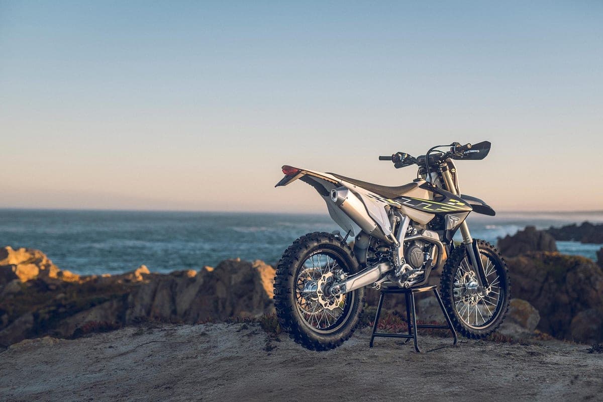 Triumph TF 250-E parked on a cliffside stand at dusk, ocean backdrop lighting up the silver bodywork and yellow TF decals