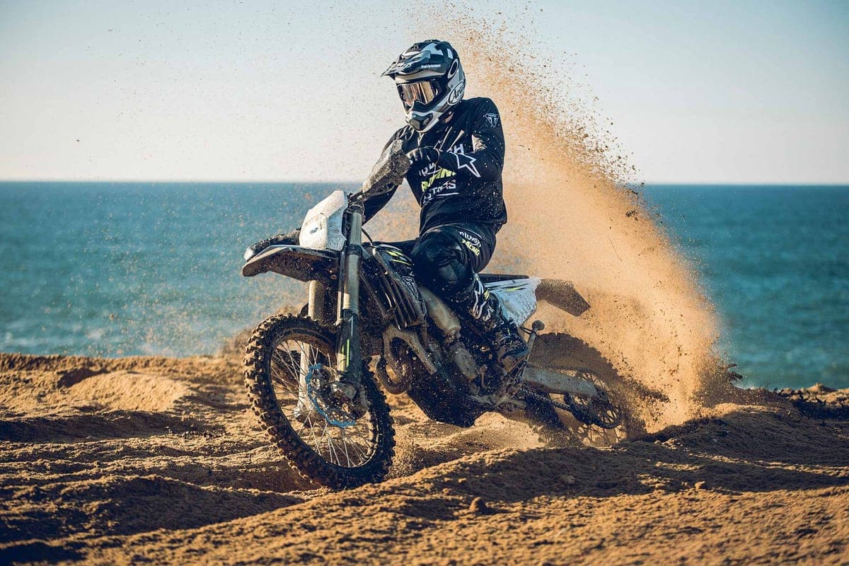 Rider in dark gear blasting the Triumph TF 250-E across a coastal sand dune, kicking up a wide roost of dirt against the sea