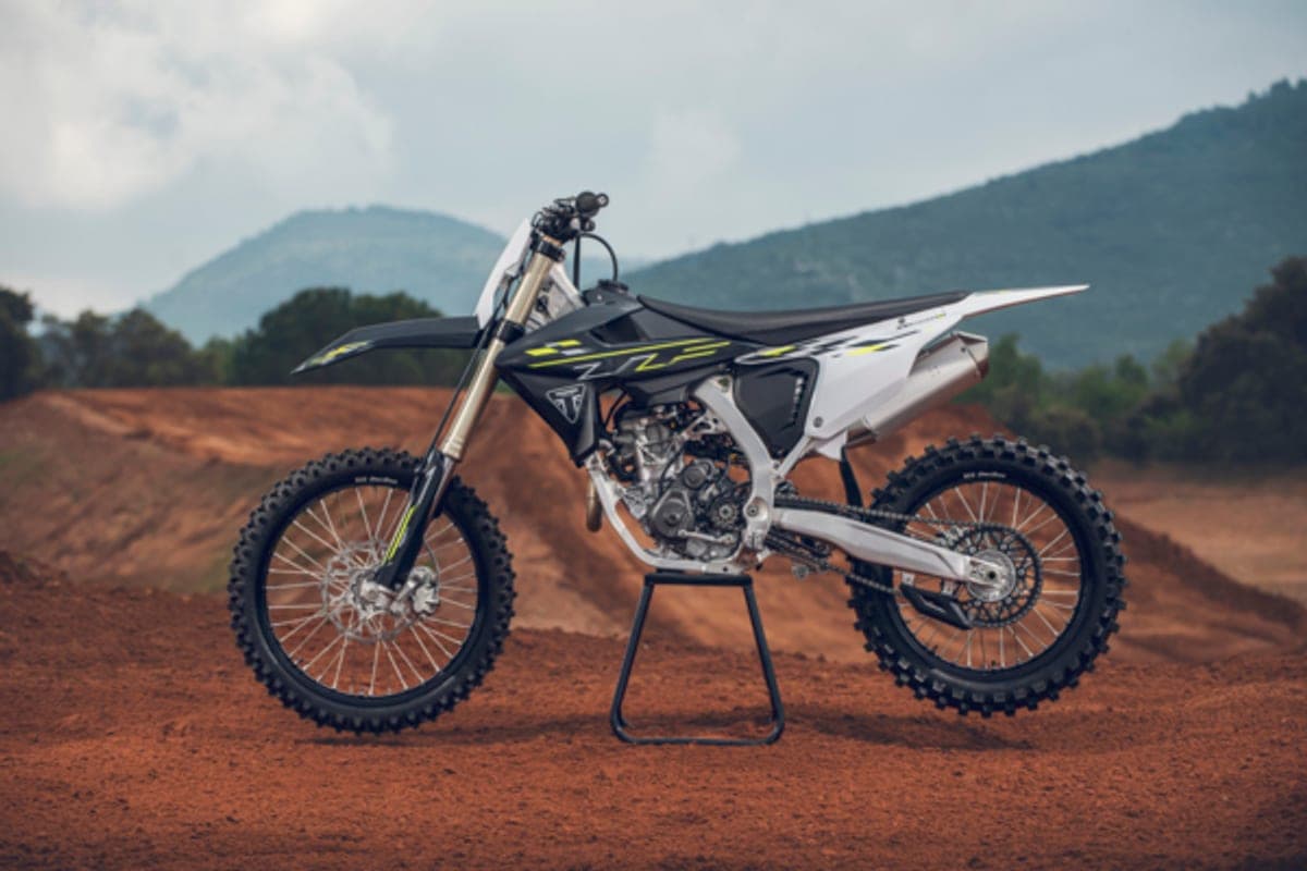 Triumph TF 250-X on location at a dirt track with red clay berms and hazy mountains behind it, side view on a work stand showing the engine and spoked wheels