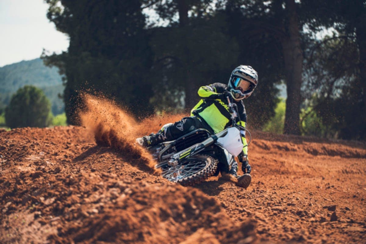 Rider in black and yellow gear leaning a white and black Triumph TF 250-X hard into a red-dirt berm, throwing a large roost of soil