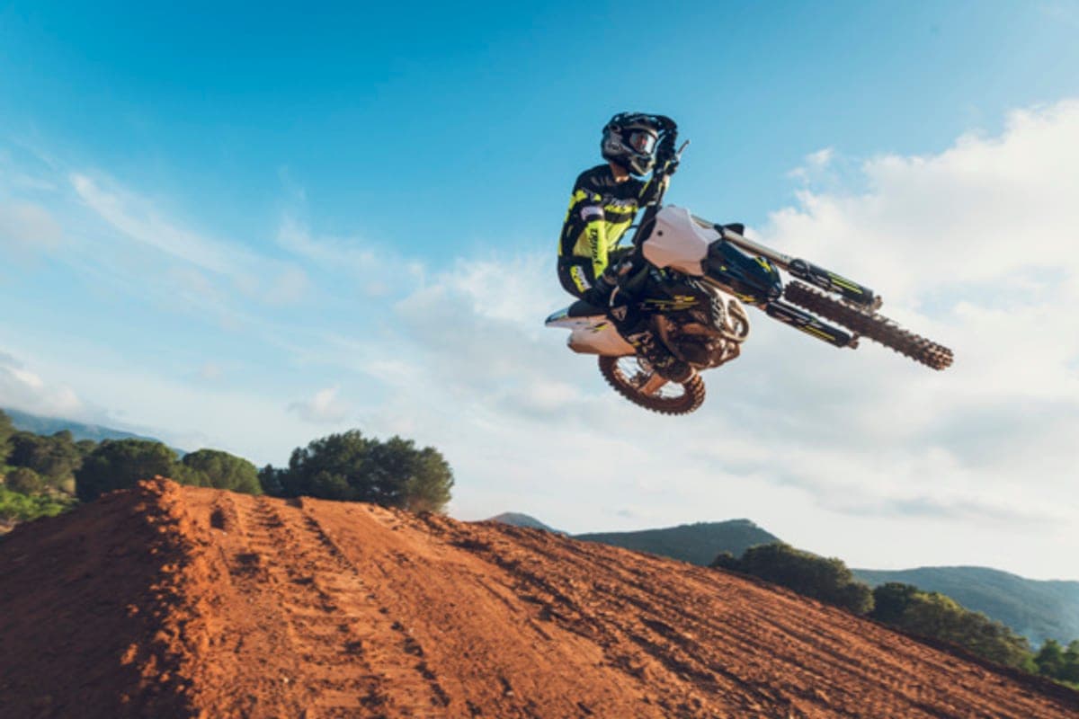 Rider in yellow and black gear scrubbing a white Triumph TF 250-X sideways off a dirt jump high above a red motocross track under a blue sky