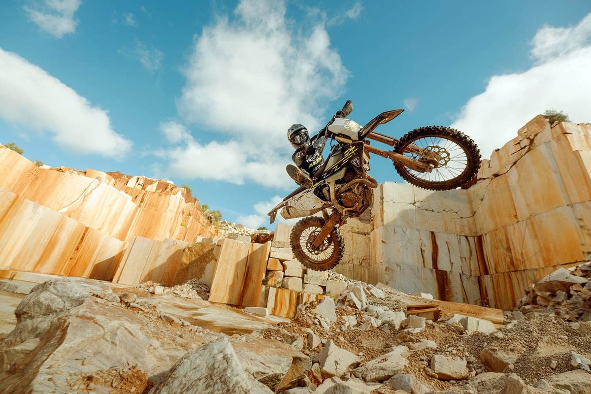 Rider in dark gear on a muddy Triumph TF 450-E launching off a rock ledge in a quarry, front wheel lofted against a blue sky