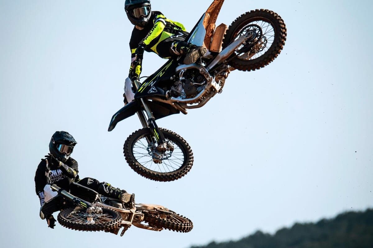 Rider in black and neon-yellow gear whipping a Triumph TF 450-X sideways mid-air above another rider on a black Triumph, blue sky backdrop