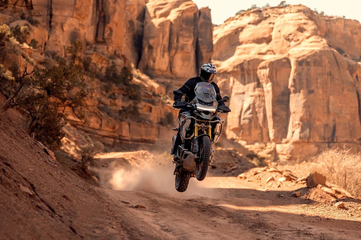 Rider in black gear catching air on a Triumph Tiger 1200 Rally Pro on a dusty desert trail flanked by red sandstone canyon walls