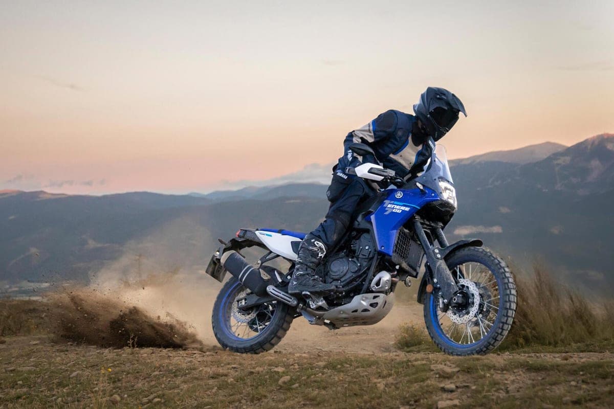 Rider in dark blue adventure gear powering a Yamaha Ténéré 700 up a dusty dirt trail at dusk, kicking up a cloud of dirt behind the rear wheel