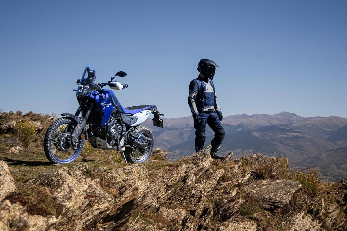 Blue and white Yamaha Ténéré 700 parked on a rocky ridge beside a standing rider in touring gear, mountain range stretching behind